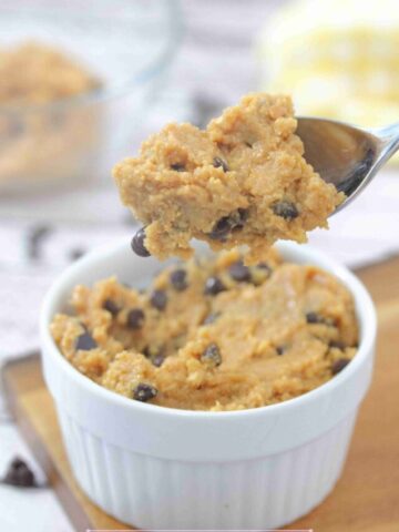 Spoonful of chocolate chip protein cookie dough being pulled out of a white ramekin that's on a wooden cutting board.