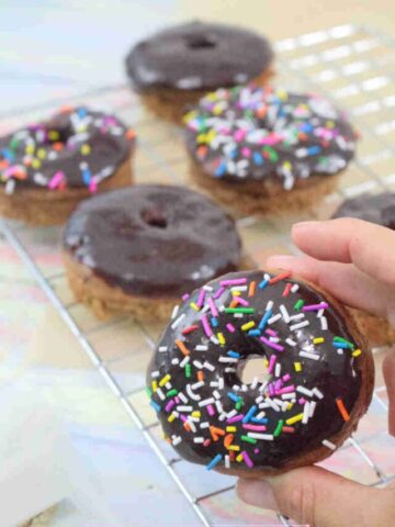 Two fingers holding up a chocolate glazed protein donut with sprinkles with more donuts behind it.