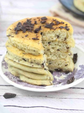 Protein pancakes with chocolate chips stacked on a white and purple platye with a slice cut out of the stack.
