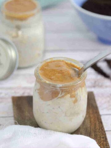 Mason jar with espresso overnight oats topped with almond butter and a spoon in it on a wooden coaster with another jar of oats and a bowl of coffee behind it.