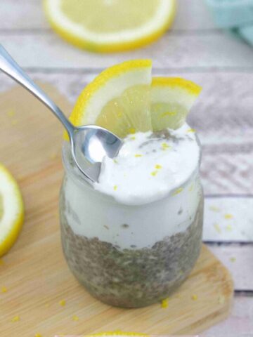 Lemon chia protein pudding topped with coconut cream in a small jar with lemons on top and a spoon in the pudding.