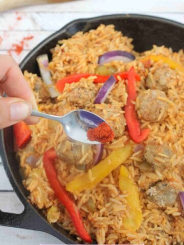 Black skillet with rice, sausages, and peppers in a tomato sauce. and a spoon of paprika being added.