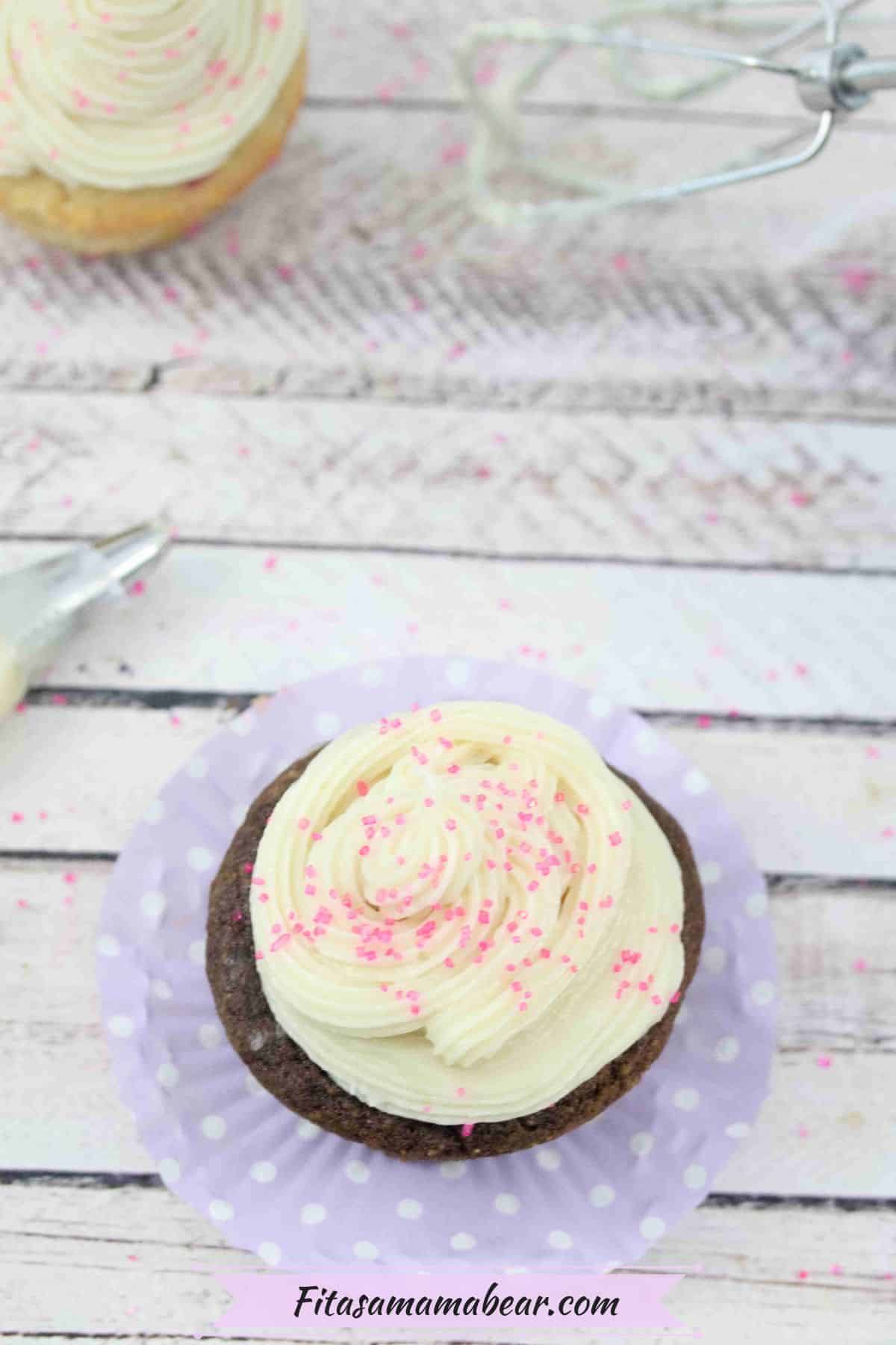 Chocolate cupcake with dairy-free buttercream frosting on a purple cupcake liner with a piping tool and more cupcake behind it.