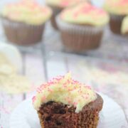 Chocolate protein cupcake with buttercream frosting with a bite taken out of it on a white chupcake liner with more gluten-free cupcakes on a cooling rack behind it.