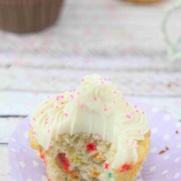 Protein cupcake with dairy-free buttercream frosting on a purple muffin liner with a bite out of the cupcake and more cupcakes behind it.