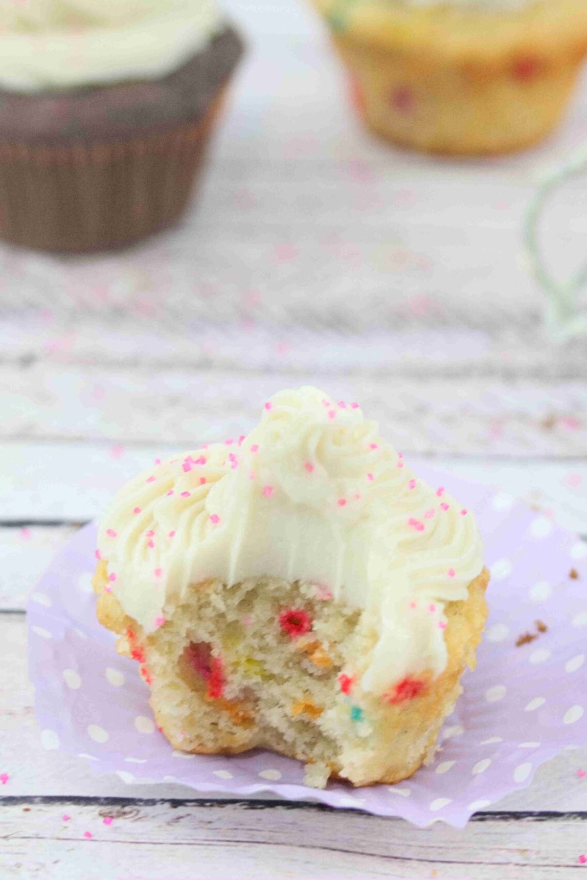 Protein cupcake with dairy-free buttercream frosting on a purple muffin liner with a bite out of the cupcake and more cupcakes behind it.