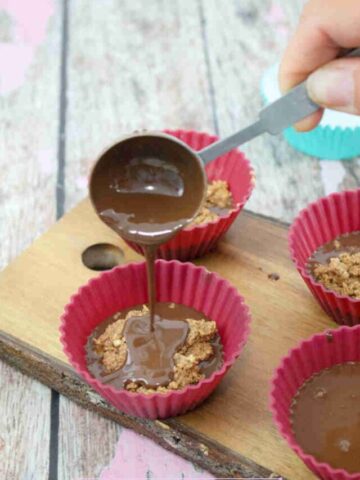 Red silicone molds on a wooden cutting board with melted chocolate being poured over almond butter.