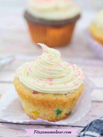 Close up of a cupcake with dairy-free buttercream frosting on top and pink shimmer sprinkles with more cupcakes behind it.