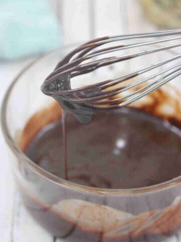 Dairy-free chocolate ganache dripping off a whisk over a glass bowl with more ganache in it.