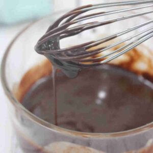 Dairy-free chocolate ganache dripping off a whisk over a glass bowl with more ganache in it.