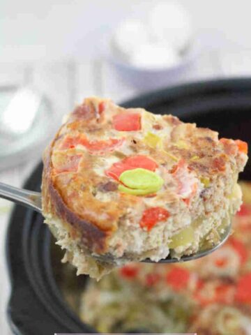 Close up of a slice of egg casserole being held over a black slow cooker of breakfast casserole.
