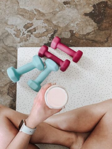 Woman sitting cross legged and holding a protein shake with small dumbbells around her.