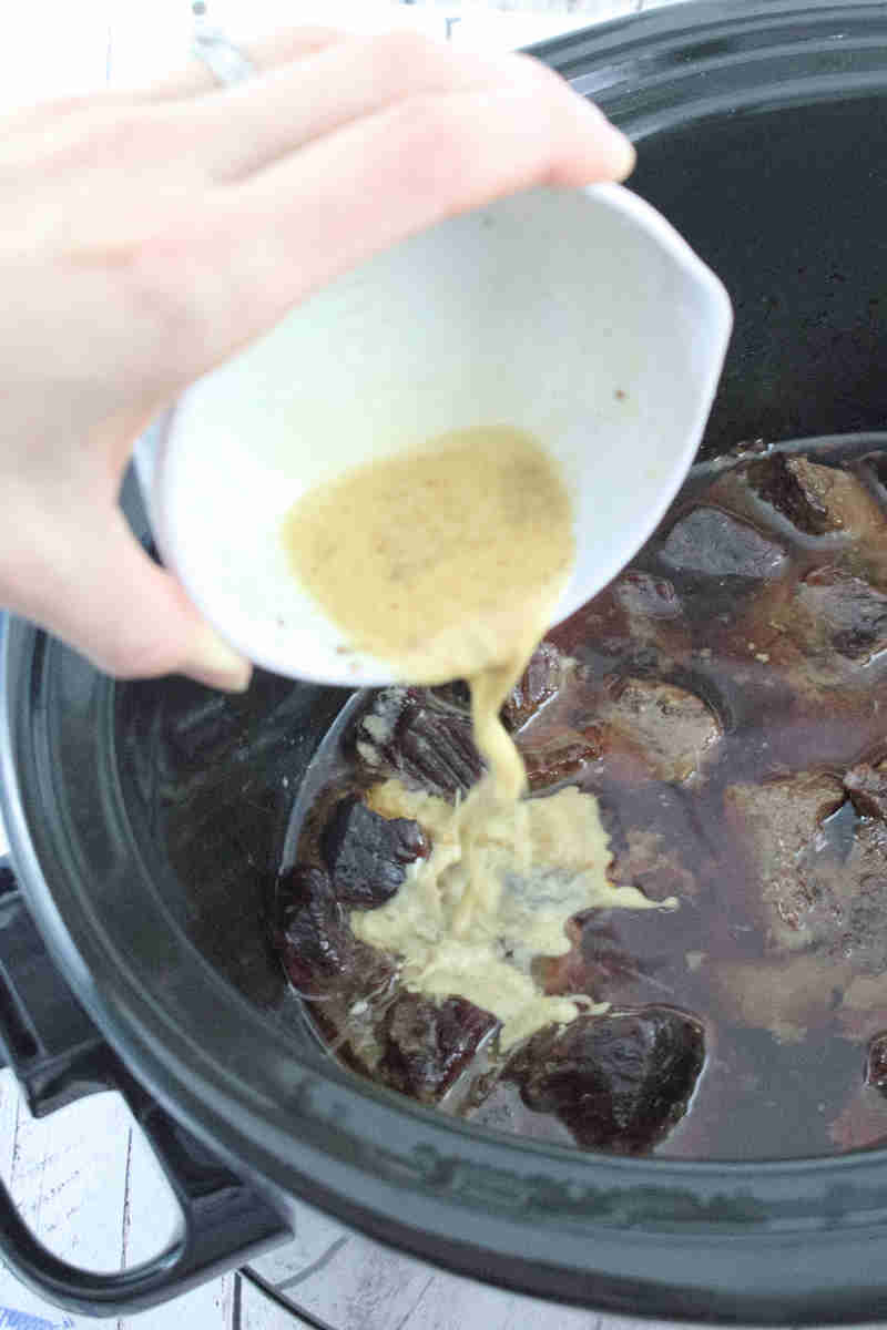 Thickening sauce being poured from a white ramekin into a black slow cooker.