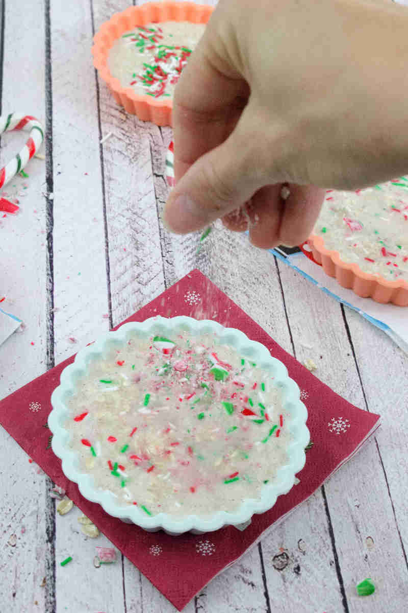 Three silicone molds with blended protein oatmeal in them in Christmas napkins and a hand sprinkling candy canes into the molds.