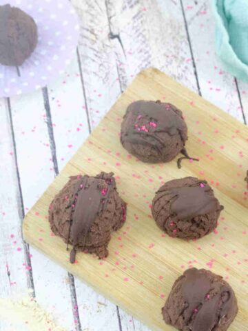 Multiple chocolate protein truffles with pink sprinkles on top on a wooden cutting board with more truffles and around it.