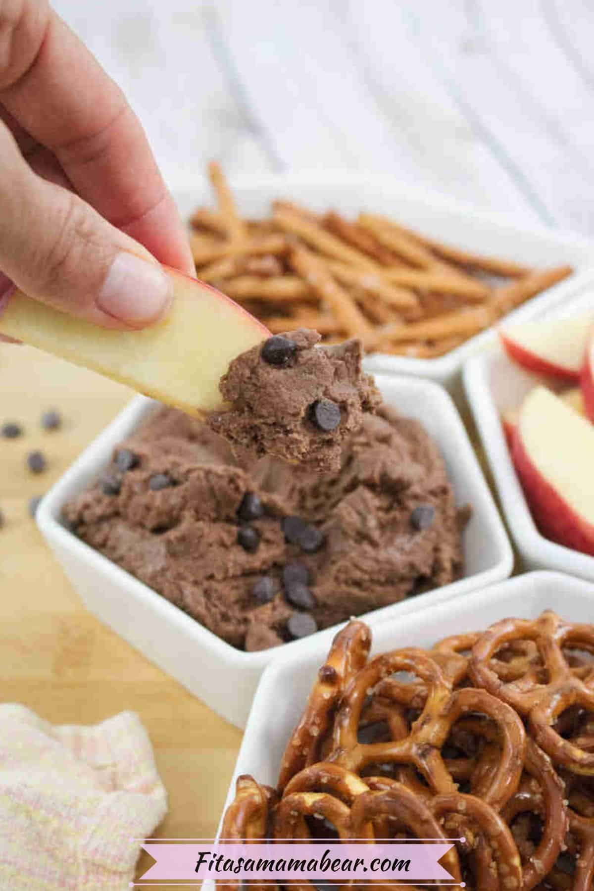 An apple slice being dipped into high protein edible brownie batter topped with chocolate chips in a white ramekin with more pretzels and apple slices around it.