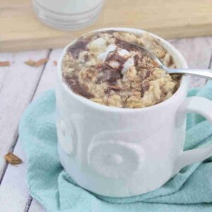 White mug with egg white oatmeal in it topped a cinnamon glaze and a spoon in the mug on a blue linen.