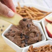 An apple slice being dipped into high protein edible brownie batter topped with chocolate chips in a white ramekin with more pretzels and apple slices around it.