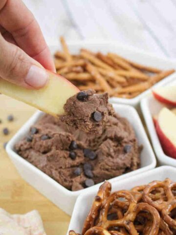 An apple slice being dipped into high protein edible brownie batter topped with chocolate chips in a white ramekin with more pretzels and apple slices around it.