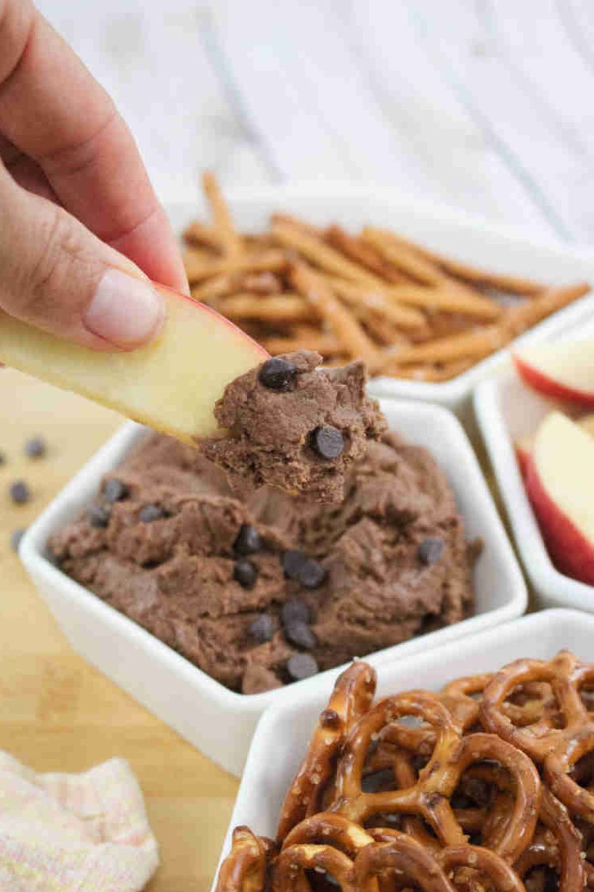 An apple slice being dipped into high protein edible brownie batter topped with chocolate chips in a white ramekin with more pretzels and apple slices around it.