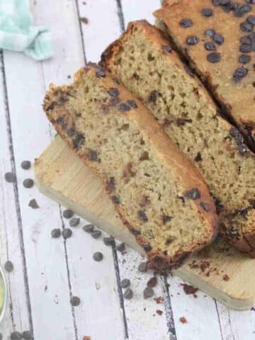 Sliced high protein banana bread with chocolate chips on a wooden cutting board with a blue linen beside it.