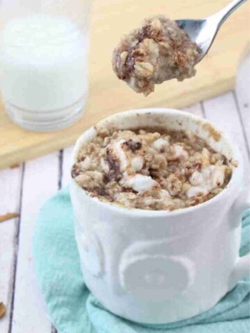 Spoon being pulled out of a White mug with egg white oatmeal in it topped a cinnamon glaze and a spoon in the mug on a blue linen.