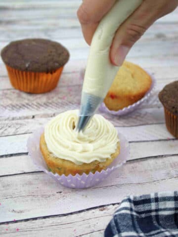 Dairy-free buttercream frosting being piped onto a vanilla protein cupcake in a purple cupcake liner.