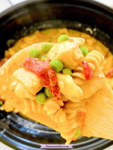 Close up of a wooden spoon with slow cooker chicken alfredo with noodles, peas, chicken and sundried tomatoes on the spoon and the black slow cooker in the background