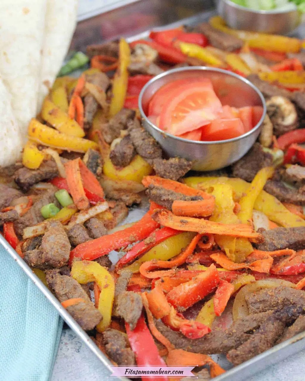 Baking tray with cooked steak, bell peppers, and veggies with a steel bowl of fresh tomatoes in the middle and flour tortillas folded to the side
