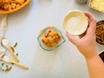Hand holding a white ramekin about to pour into a plastic baggie of raw chicken with more ingredients for pasta around it.