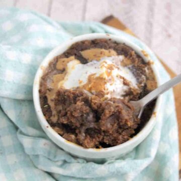 Chocolate protein pwoder mug cake topped with peanut butter and coconut whippe cream in a white mug on a blue linen.