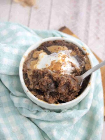 Chocolate protein pwoder mug cake topped with peanut butter and coconut whippe cream in a white mug on a blue linen.