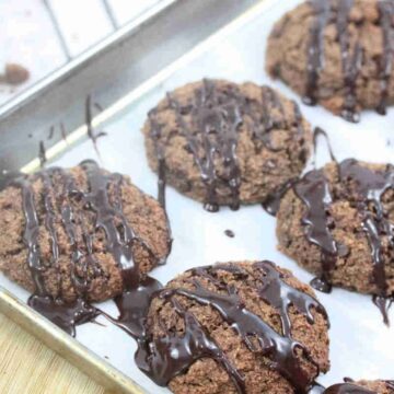 Chocolate almond flour cookies drizzled with chocolate on a piece of parchment paper on a baking sheet.