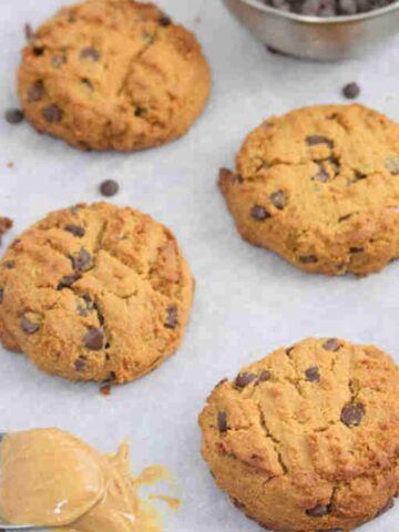 Peanut butter chocolate chip protein cookies on a parchment paper lined baking tray with a spoon of peanut butter on the tray too.