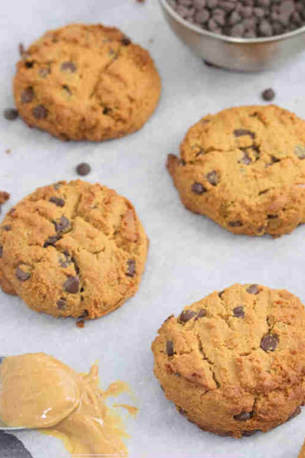 Peanut butter chocolate chip protein cookies on a parchment paper lined baking tray with a spoon of peanut butter on the tray too.
