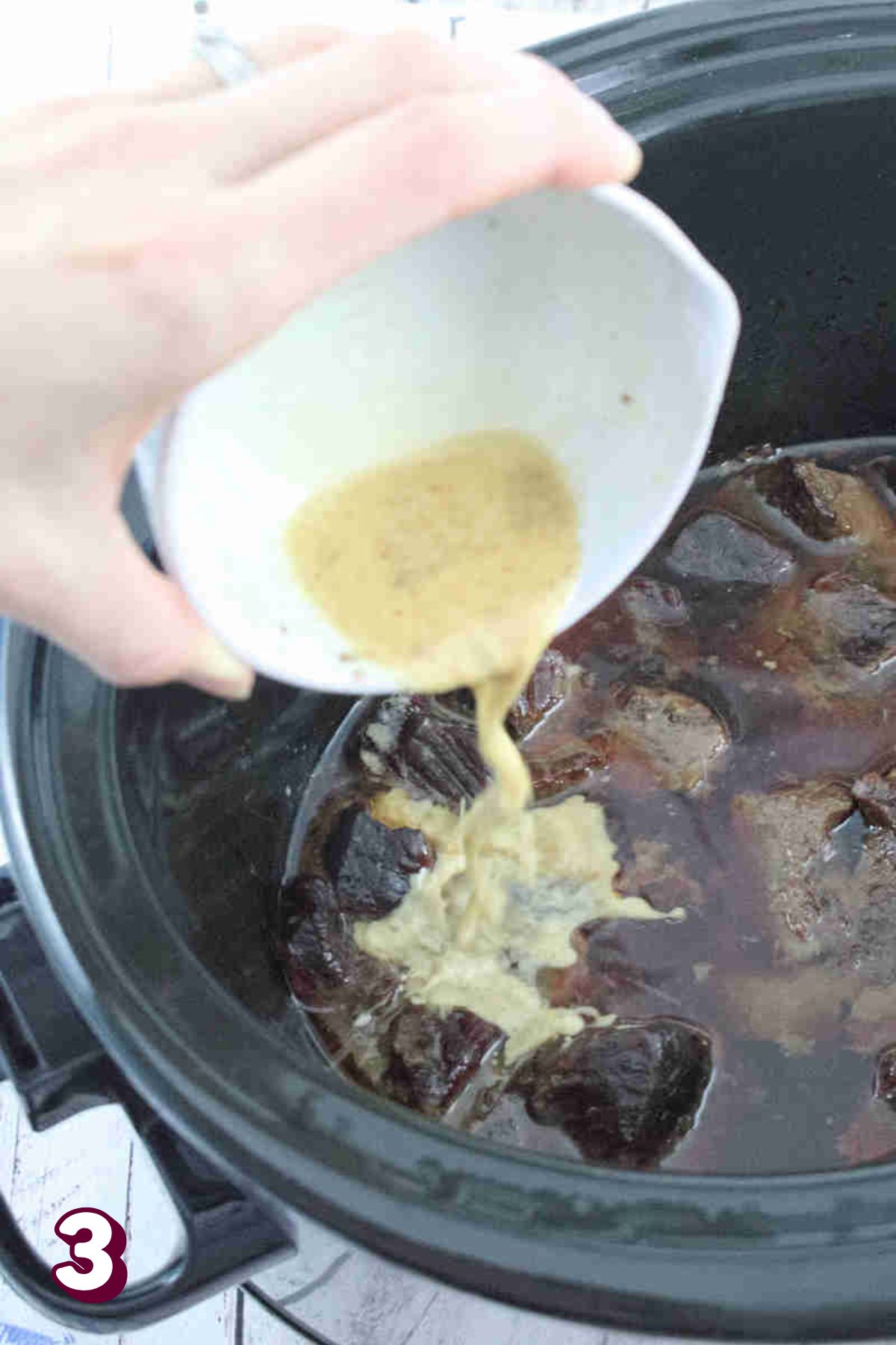 Thickening sauce being poured from a white ramekin into a black slow cooker.