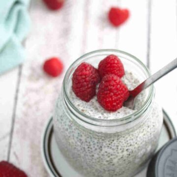 Keto chia pudding in a mason jar with raspberries on top and a spoon in the jar on a white plate with berries and a blue linen behind it.