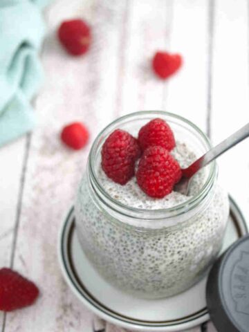 Keto chia pudding in a mason jar with raspberries on top and a spoon in the jar on a white plate with berries and a blue linen behind it.