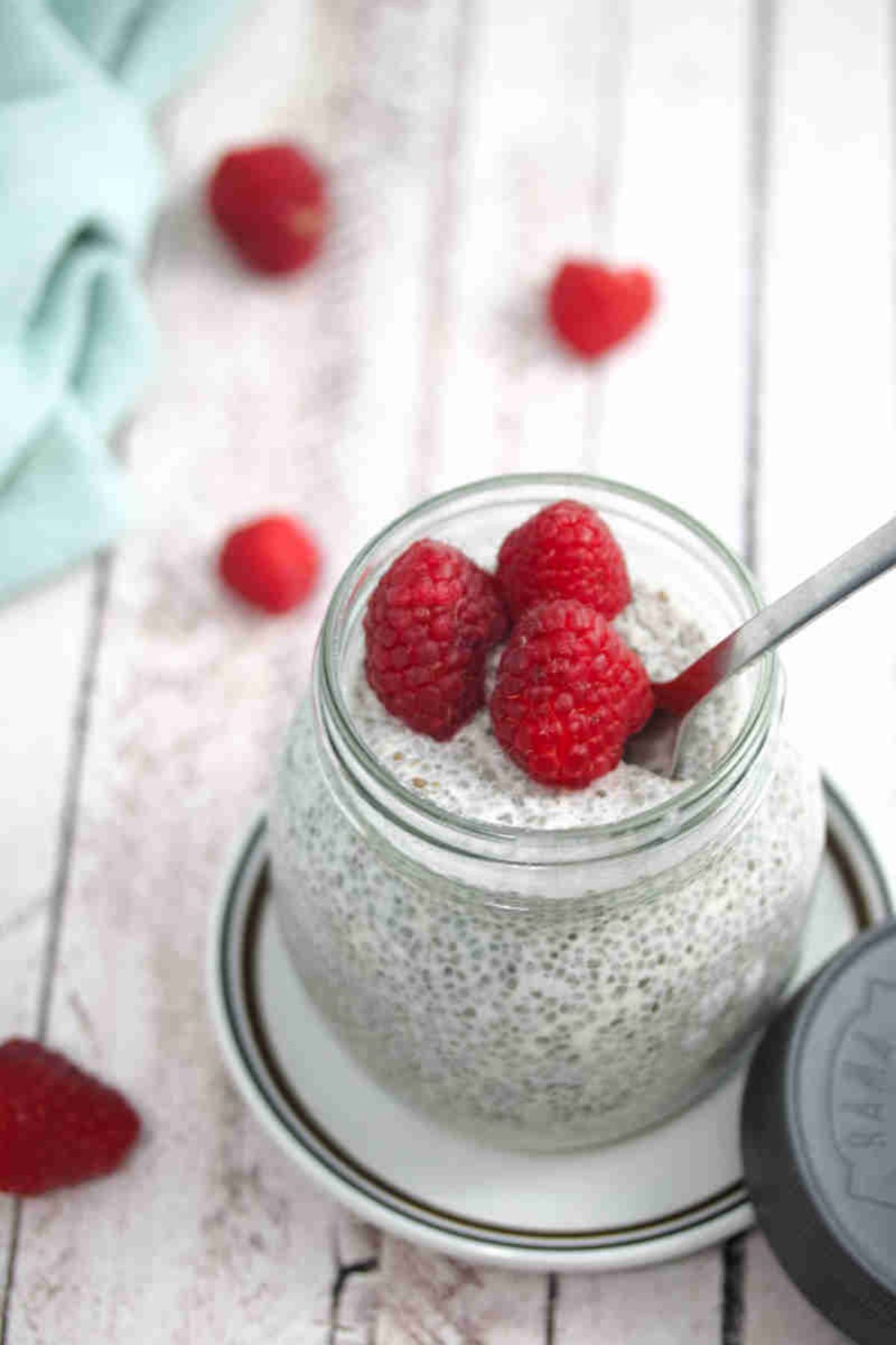 Keto chia pudding in a mason jar with raspberries on top and a spoon in the jar on a white plate with berries and a blue linen behind it.