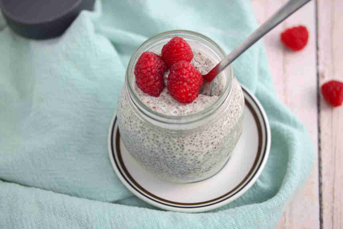 Keto chia pudding in a mason jar with raspberries and a spoon in the jar on a white plate which is on a blue linen.