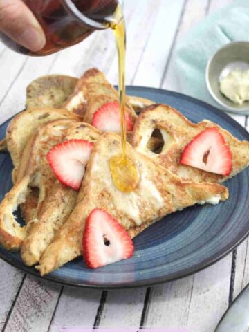 A blue plate with protein french toast in triangle slices with strawberries on it and maple syrup being poured on top.
