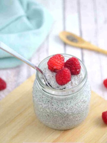 Keto chia pudding in a mason jar with raspberries on top and a spoon in the jar on a wooden cutting board with berries and a blue linen behind it.