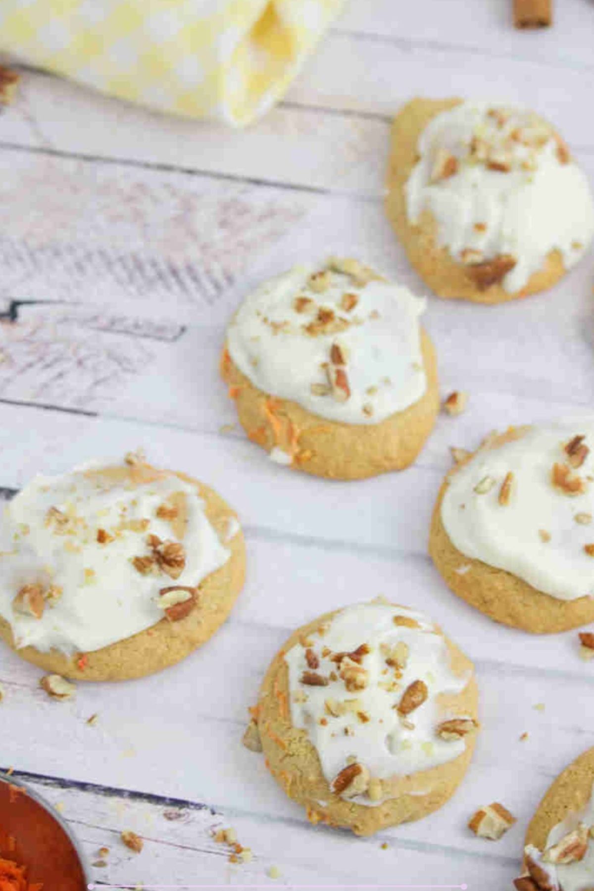 Dairy-free collagen carrot cookies topped with cream cheese frosting and pecans on a piece of parchment paper with shredded carrots beside them.