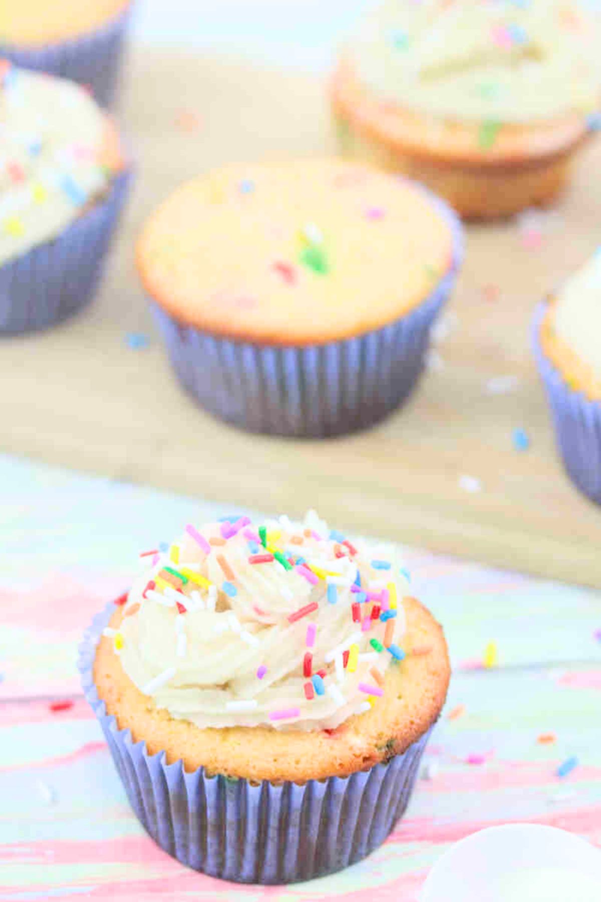 Vanilla protein cupcake with dairy-free frosting and sprinkles in a blue cupcake liner with more cupcakes behind it on a cutting board.