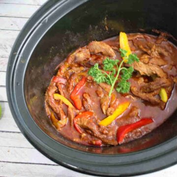 Black slow cooker with beef fajitas and bell peppers in it in a tomato sauce with ingredients around the slow cooker.