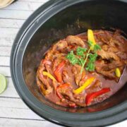 Black slow cooker with beef fajitas and bell peppers in it in a tomato sauce with ingredients around the slow cooker.