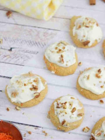 Dairy-free collagen carrot cookies topped with cream cheese frosting and pecans on a piece of parchment paper with shredded carrots beside them.