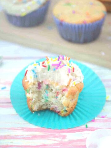 Vanilla protein cupcake with frosting and sprinkles on a blue cupcake liner with more cupcakes behind it.