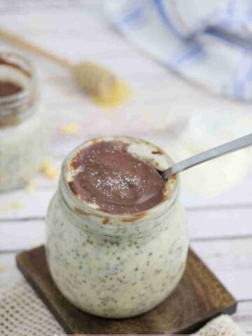 Mason jar with overnight protein oats topped with chocolate on a wooden coasted with a spoon, nuts and another jar around it.