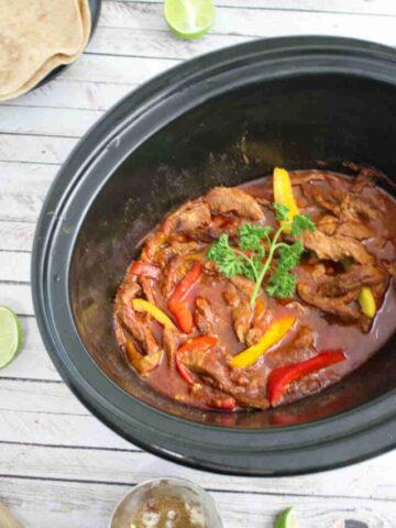 Black slow cooker with beef fajitas and bell peppers in it in a tomato sauce with ingredients around the slow cooker.